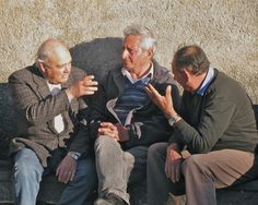 three men sitting on a bench talking to each other and one is holding his hand out