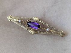 "This enchanting antique brooch is made from solid 14k gold and features a faceted amethyst stone and two genuine seed pearls on either side, set in a detailed geometric art s-and-crafts style setting. The back has an early style locking c-clasp closure and is stamped \"14k\" for metal content along with a maker's mark. It weighs 4.32g, measures approx 1 7/8\" x 5/8\" and is in very good antique condition." Lavender Brooch Jewelry As A Gift, Elegant Purple Gemstone Brooches, Elegant Amethyst Brooches For Wedding, Purple Amethyst Gemstone Brooches, Antique Hallmarked Purple Brooches, Antique Purple Hallmarked Brooches, Amethyst Brooch Jewelry Gift, Purple Amethyst Brooches For Wedding, Purple Gemstone Brooches For Weddings