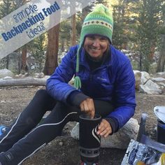 a smiling woman sitting on the ground next to a campfire with food and water