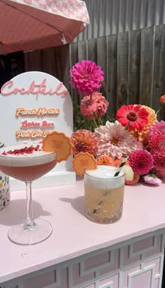 a table topped with two cocktail glasses filled with liquid next to flowers and an umbrella