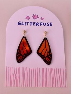 a pair of orange and black butterfly shaped earrings on top of a pink card with the words glitterfuse