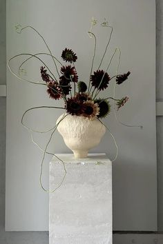 a white vase filled with lots of flowers on top of a block of concrete in front of a wall