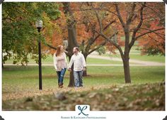 a man and woman are walking through the park in front of trees with orange leaves