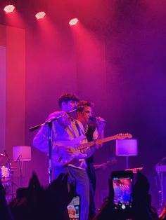 two young men are singing on stage with microphones in front of them, and one man is playing the guitar