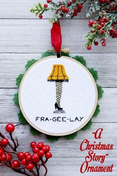 a cross stitch christmas ornament with a lamp on it and red berries in the foreground