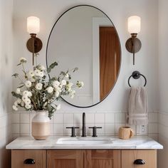 a bathroom vanity with a round mirror above it and flowers in a vase on the counter