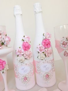 three wine bottles decorated with pink flowers and lace, sitting next to two champagne flutes