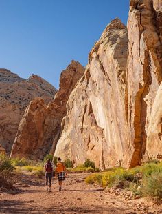 two people walking down a dirt road in the desert with large rocks on either side