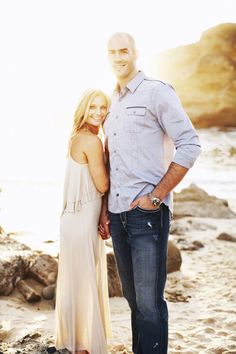 a man and woman standing next to each other on the beach in front of some rocks