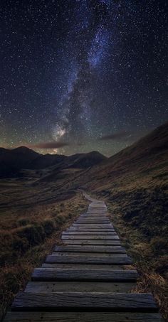 a wooden walkway leading to the stars in the night sky with mountains and grass behind it