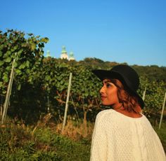 a woman wearing a black hat standing in front of a vineyard with vines behind her