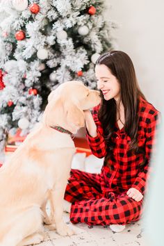 a woman in red and black pajamas petting a yellow labrador retriever near a christmas tree