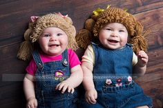 two baby girls wearing overalls and hats on top of a wooden floor, one smiling at the camera