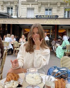 a woman covering her mouth while sitting at a table with pastries on it in front of a cafe