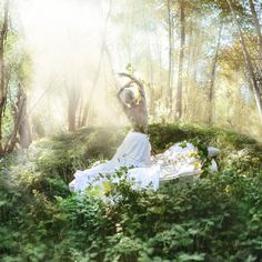 a woman sitting in the middle of a forest surrounded by green plants and trees, with her hair blowing in the wind