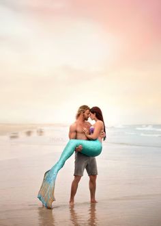 a man holding a mermaid tail while standing next to a woman on the beach at sunset