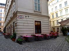 an old building with tables and chairs on the sidewalk in front of it, surrounded by tall buildings