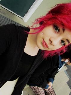 a girl with red hair and piercings on her nose looking at the camera while standing in front of a building