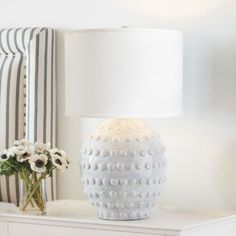 a white table lamp sitting on top of a dresser next to a vase with flowers