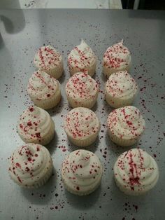 twelve cupcakes with white frosting and red sprinkles on a silver surface