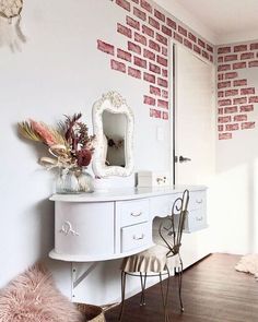 a white desk with a mirror and some flowers on it in front of a brick wall