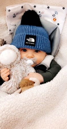 a baby in a bed holding a stuffed animal and wearing a blue beanie hat