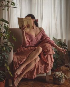 a woman sitting in a chair reading a book with her legs crossed and wearing a pink dress