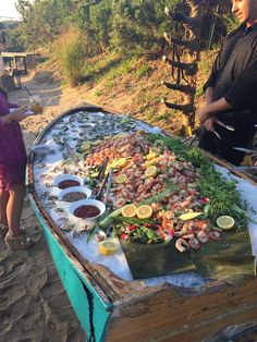 a man and woman standing in front of a large platter of food on the beach