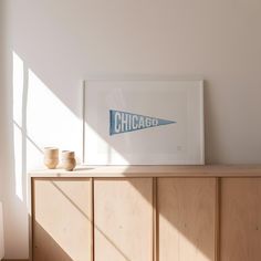 a framed chicago sign sitting on top of a wooden cabinet next to two cups and a vase