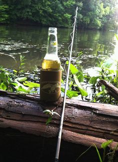 a beer bottle sitting on top of a log next to a fishing pole in the water