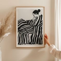 a woman is holding up a black and white painting on the wall next to a plant