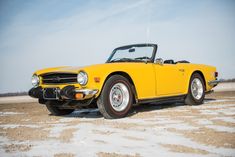 an old yellow sports car is parked on the beach in front of some sand and blue sky