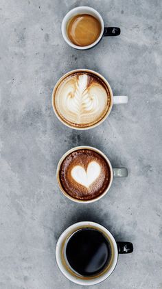 three cups of coffee with hearts in them on a gray table top next to each other