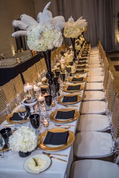 a long table is set with black and gold place settings, white flowers and candles