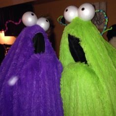 two green and purple monster costumes with white balls on their eyes, one is wearing a black hat