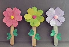 three wooden sticks with flowers on them sitting in front of a gray wall and one is holding a paper flower