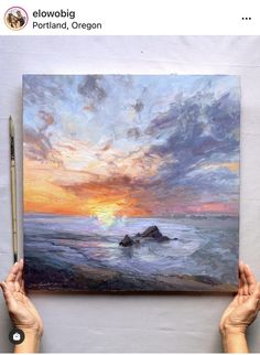 two hands holding up a painting with the sun setting over the ocean in the background