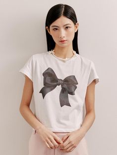 This is a short-sleeved, crewneck t-shirt that features a large bow graphic on the front. The graphic gives the impression of a textile bow tied at the center, adding a decorative and feminine touch to the classic tee. The shirt's design is simple and clean, making it versatile for various occasions. The combination of the casual cut and the eye-catching graphic creates a playful yet chic garment.- The bow graphic serves as a bold statement piece on the otherwise minimalist t-shirt.- Its classic crewneck and short-sleeve design make it a staple piece for any casual wardrobe.- The contrast between the graphic and the shirt suggests a design that's both modern and stylish. Chic Short Sleeve Graphic T-shirt, Chic Cotton Tops With Bow, Casual White Tops With Bow, Trendy White Tops With Bow Print, Cotton Tops With Bow Print And Short Sleeves, Chic Cotton Tops With Bow Detail, Trendy Short Sleeve Tops With Bow, Casual Short Sleeve Tops With Bow, Trendy White Top With Bow