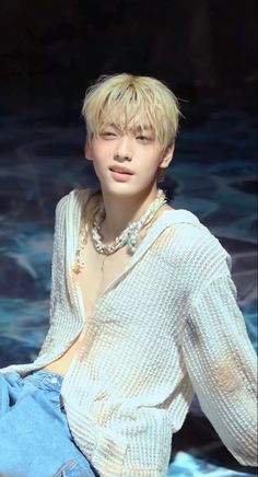 a young man with blonde hair sitting in front of a swimming pool wearing blue jeans and a white shirt