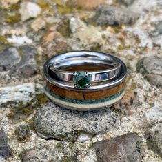 two wedding bands with a green stone in the middle on top of some rocks and gravel