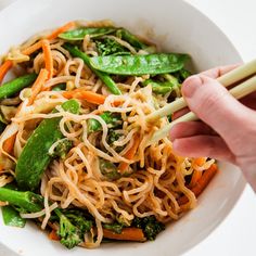 a person holding chopsticks over a bowl of noodles and vegtables