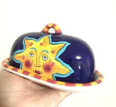 a hand holding a blue and yellow plate with a painted sun on it's lid