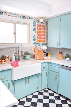 an orange cat sitting on top of a kitchen sink next to blue cabinets and counter tops