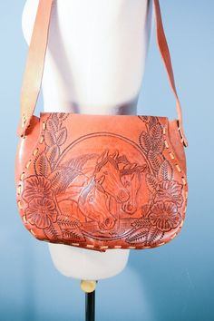 "This is a fabulous handmade hand-tooled leather shoulder bag. The leather is tooled on both sides of the bag. On the front: No Hunting and Gabi. The back has flowers and horses in great detail. There is leather stitching along the sides of the bag. A flap on the front slips into a leather strap. The bag is unlined and has no pockets. Woodstock era, late 60s early 70s. Please remember that all vintage items are at least 20 years old. Usually, they have been used and loved, there may be minor fla