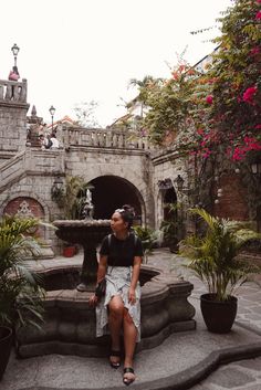 a woman sitting in front of a fountain surrounded by potted plants and other greenery