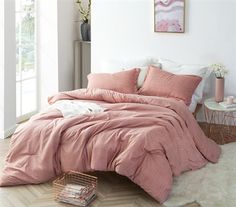 a bed with pink comforter and pillows in a white room next to a window