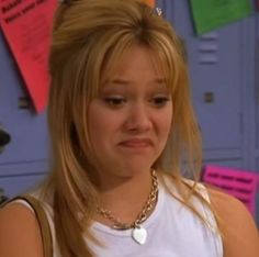 a woman with blonde hair wearing a white shirt and gold chain around her neck looking at the camera