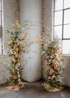 two tall floral arrangements in front of a white brick wall with windows on either side