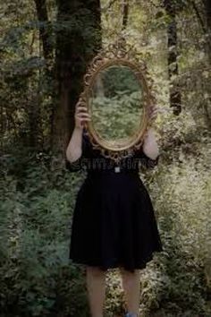 a woman in a black dress is holding a mirror over her face while standing in the woods