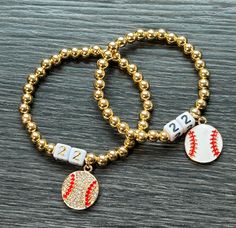 two gold beaded bracelets with baseball charms and numbers on them, sitting on a wooden surface